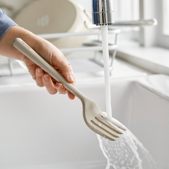 A hand washing a taupe silicone fork under running water in a kitchen sink.