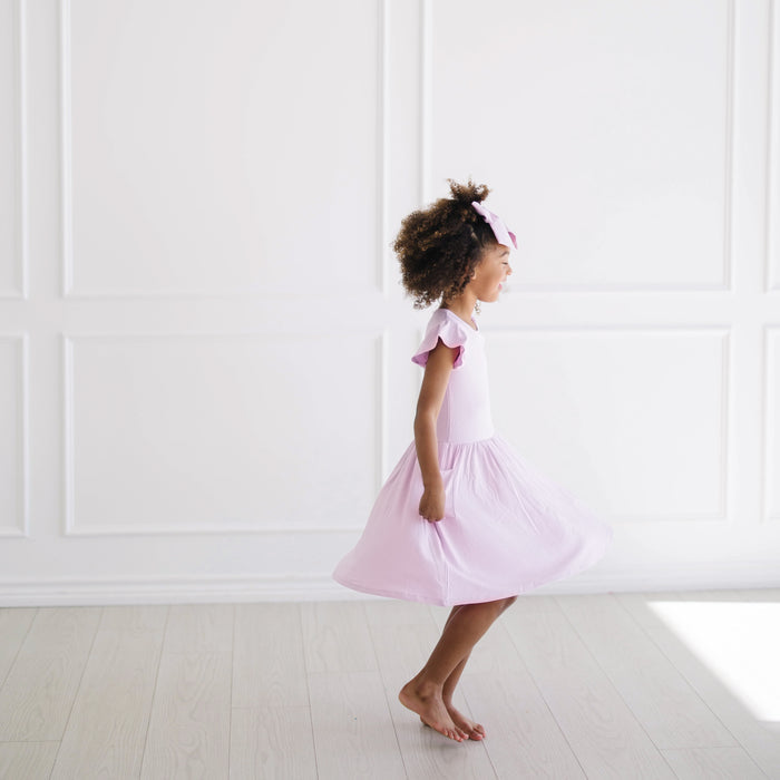 Side view of the Pocket Dress in Thistle on a young girl twirling in front of a white wall