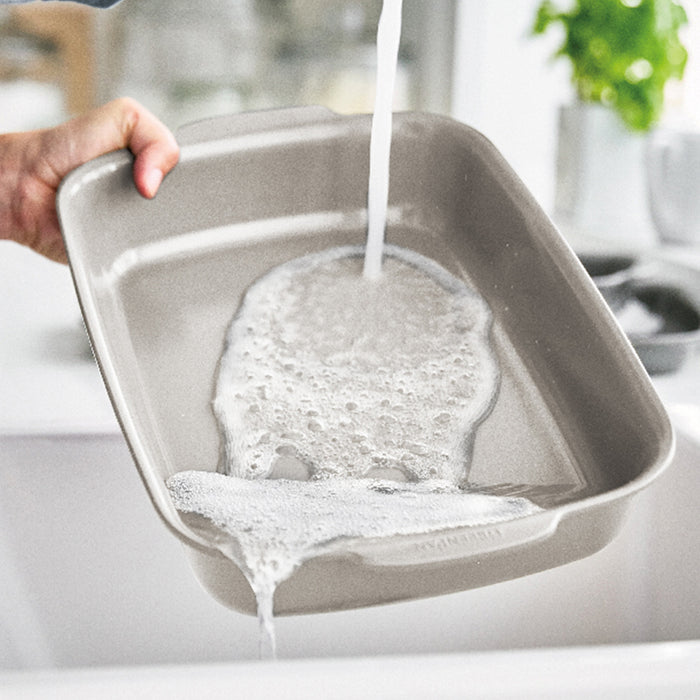 Rectangular ceramic baker being rinsed under running water, soap visible on the surface.