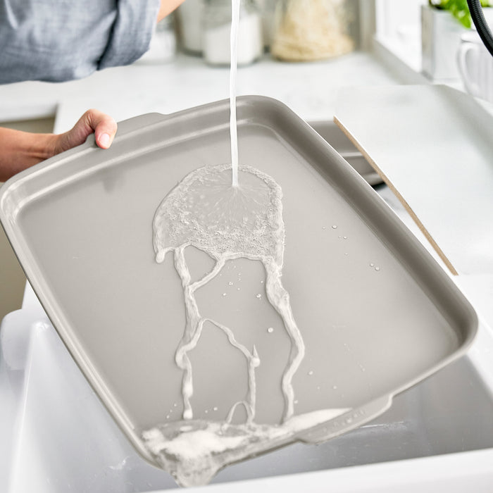 A person rinses a taupe ceramic nonstick sheet pan set under running water in a kitchen sink.
