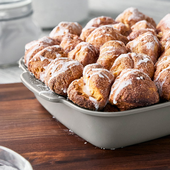 Two white ceramic nonstick cake pans holding monkey bread with icing, shown on a wooden surface. Set of 2.