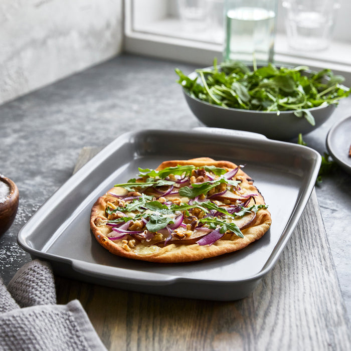 Flatbread with red onions, walnuts, and arugula on a gray ceramic nonstick pan; extra arugula visible in the background.