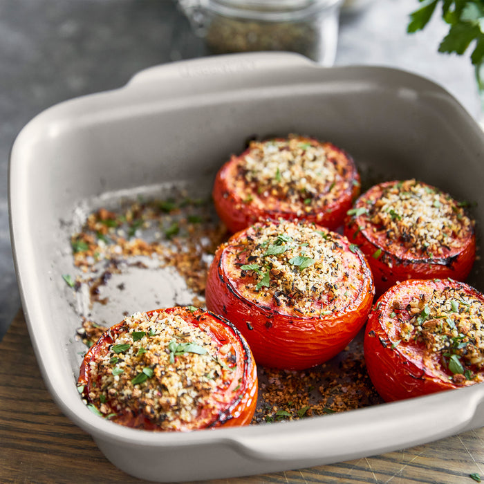 Square ceramic baker with five stuffed tomatoes topped with breadcrumbs, shown on a wooden surface.