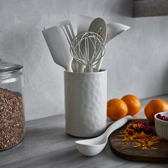 Bisque silicone utensil set with crock, eight pieces shown on a counter beside oranges, grains, and a ladle.