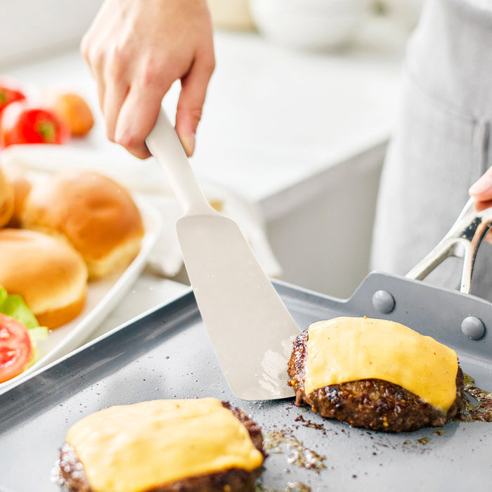 Silicone spatula from 8-piece Bisque utensil set flipping burger on griddle; buns and veggies in background.