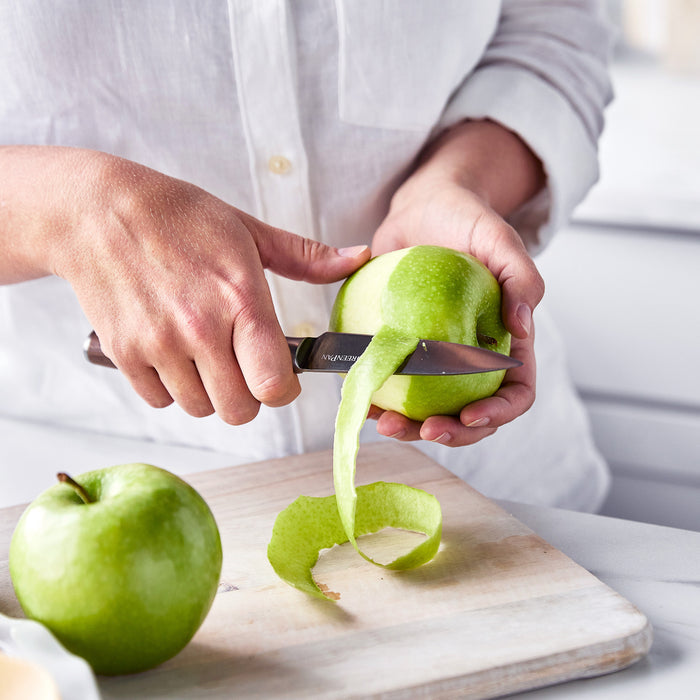 Person peeling a green apple with a 4 paring knife with a walnut handle next to a cutting board.