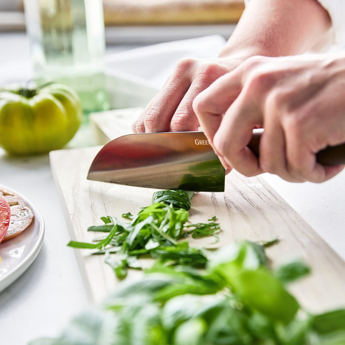 Hands chopping herbs with a small Santoku knife featuring a walnut handle; tomatoes visible in the background.