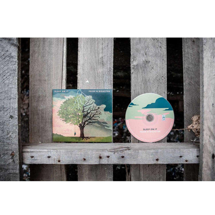 A Sleep On It "Pride & Disaster" CD and disc rest on a weathered wooden surface, displaying cover art of a tree with green and bare branches under a cloudy sky with lightning, representing the album by the band Sleep On It.