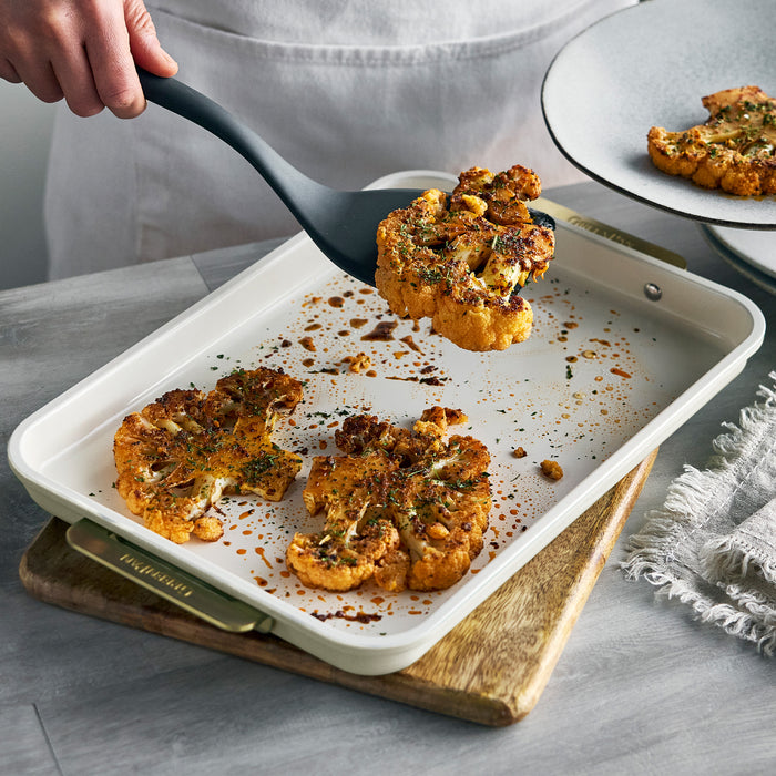 A black spatula lifts a roasted cauliflower steak from a Reserve 8-piece bakeware set onto a plate.