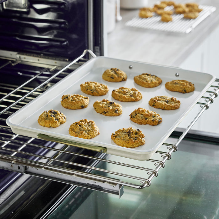 Cream 18-piece cookware and bakeware set with fresh cookies on a baking tray, more cooling in the background.