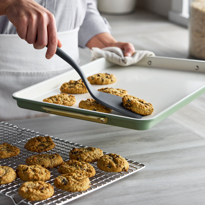 Cookies being transferred with a spatula from a sage green bakeware pan to a cooling rack.
