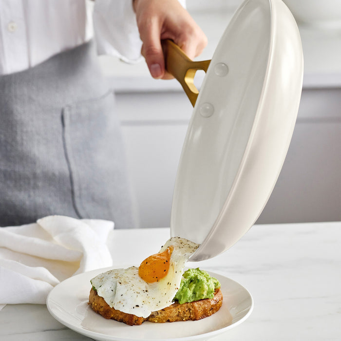Person pouring a cream frypan with a gold handle onto a plate with avocado and an egg.
