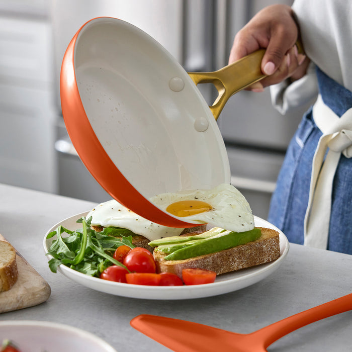 Person using a dark orange frying pan with a gold handle to place an egg on a plate of toast and vegetables.