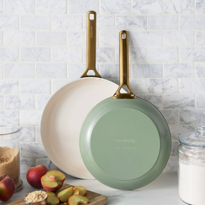 Two green frying pans on a kitchen counter with apples and a cutting board.