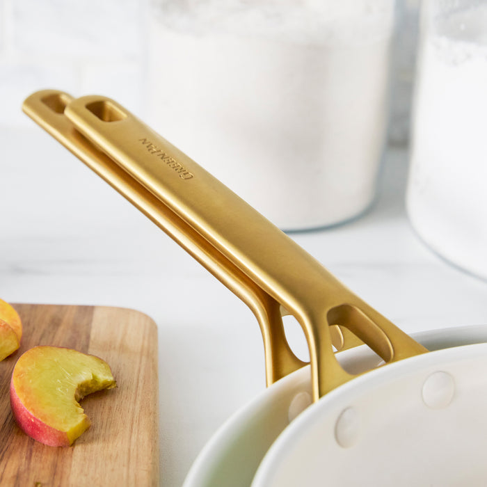 Light blue frypans with gold handles on a white surface with apples and a cutting board.