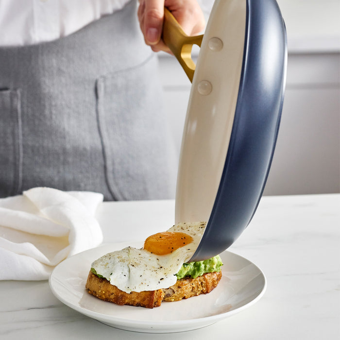 Person using a dark blue frying pan to place an egg on toast on a white plate.