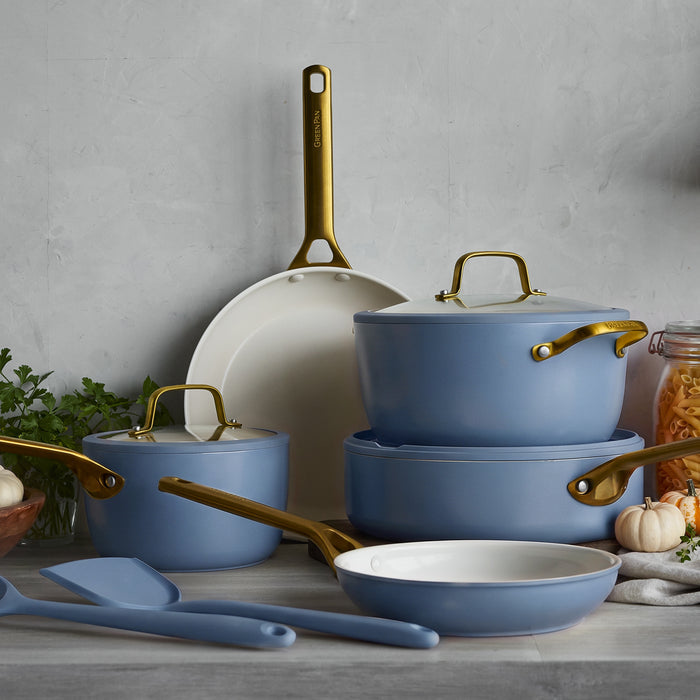 Blue 10-piece ceramic cookware set with gold handles, arranged on a counter beside utensils and fresh herbs.