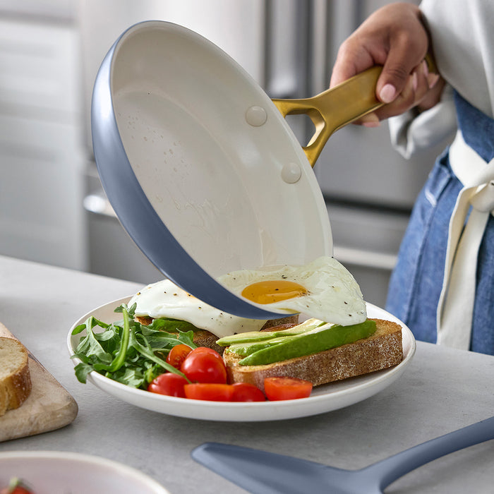 Person holding a blue frying pan with a fried egg and avocado on toast, over a plate of salad.