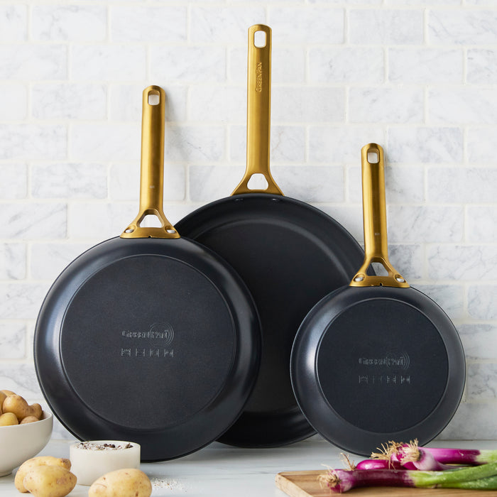 Set of three black frying pans with gold handles on a white tiled wall background
