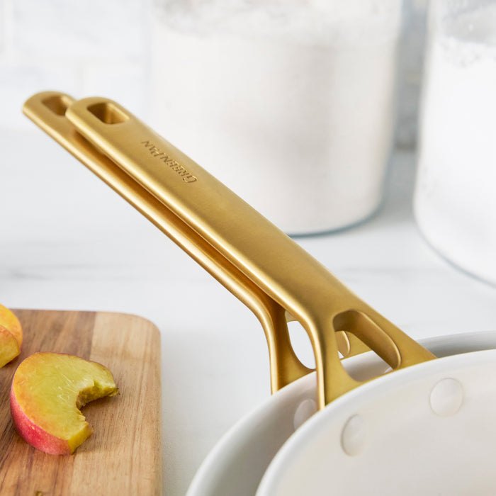 Taupe gray frypans with gold handles on a white surface with apples and a cutting board in the background