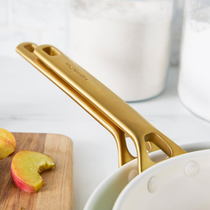 Gold frypan handles on a white surface with apples and a cutting board