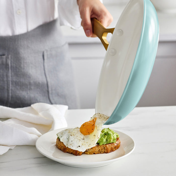 Person using a light blue frying pan to place an egg on a piece of toast with avocado on a white plate.