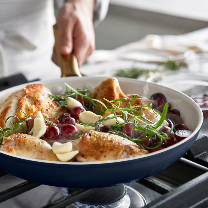 Dark blue frying pan with roasted chicken, grapes, and herbs on a stove