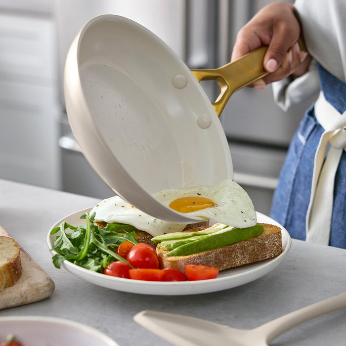 Person using a taupe frying pan with gold handle to place an egg on a plate with avocado and tomatoes.