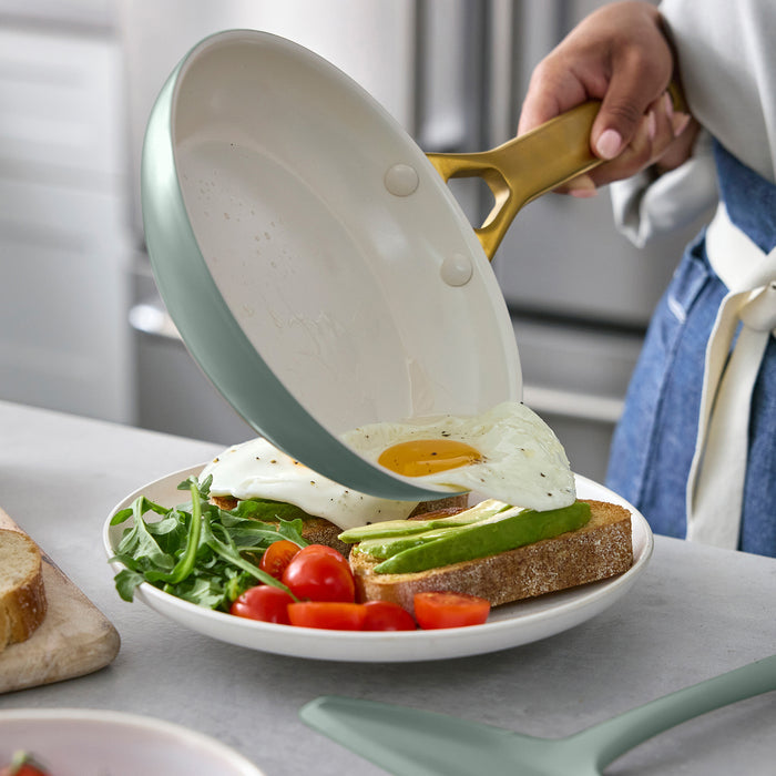 Person holding a green frying pan with eggs over a plate of toast, avocado, and vegetables.