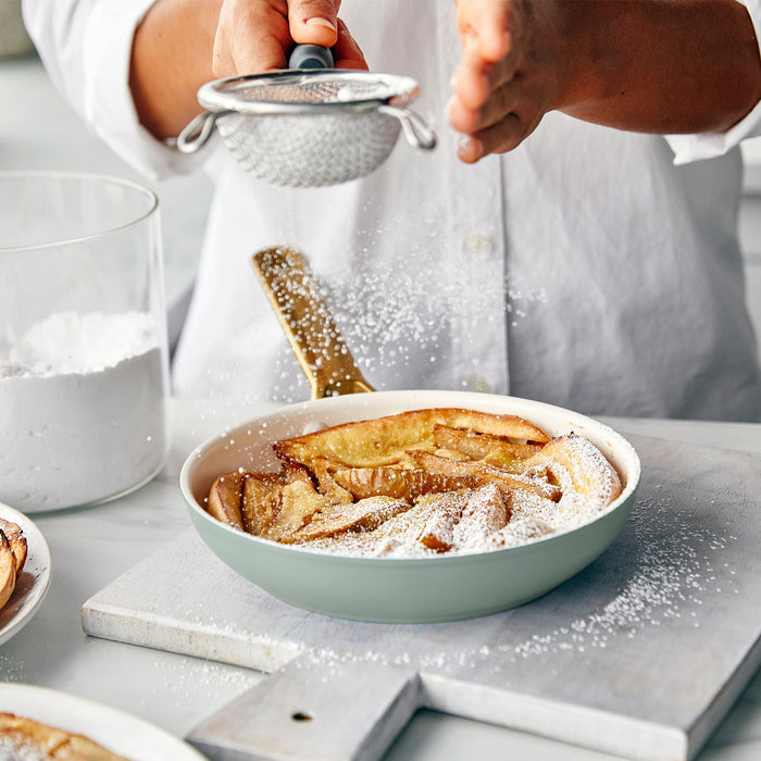 Person dusting powdered sugar over a dish of baked dessert on a marble surface.