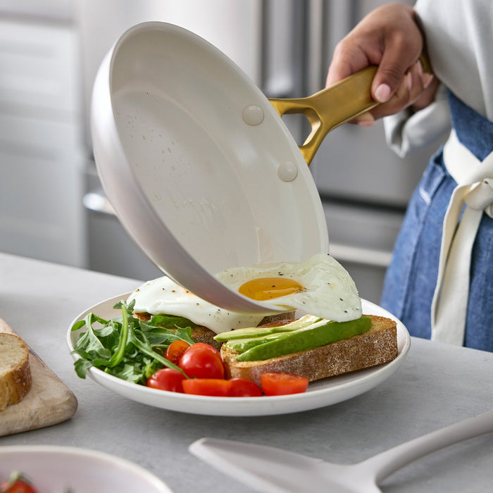 Person holding a taupe gray frying pan with a fried egg over a plate of toast, avocado, and vegetables.