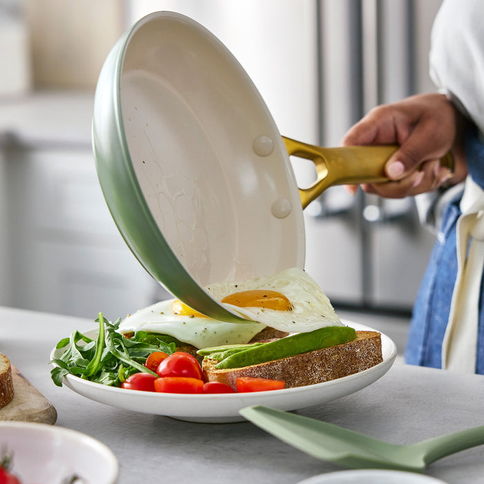 Person holding a sage green frying pan over a plate with a sandwich and vegetables