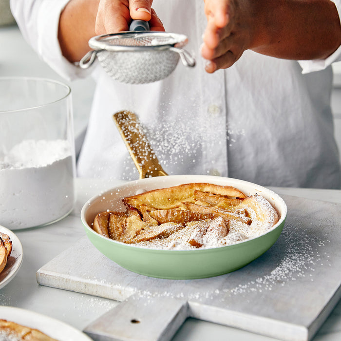Person dusting a dish of apple tart with powdered sugar in a kitchen setting.