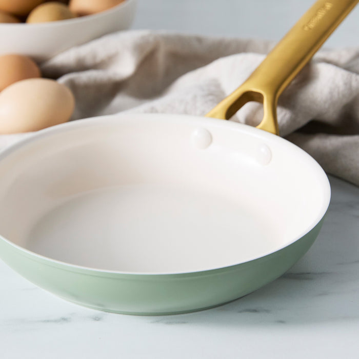Sage green frying pan with a gold handle on a marble surface, eggs in the background.