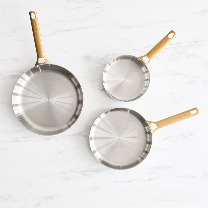 Three stainless steel frypans with gold handles on a white marble surface, in 8, 10, and 12 sizes.