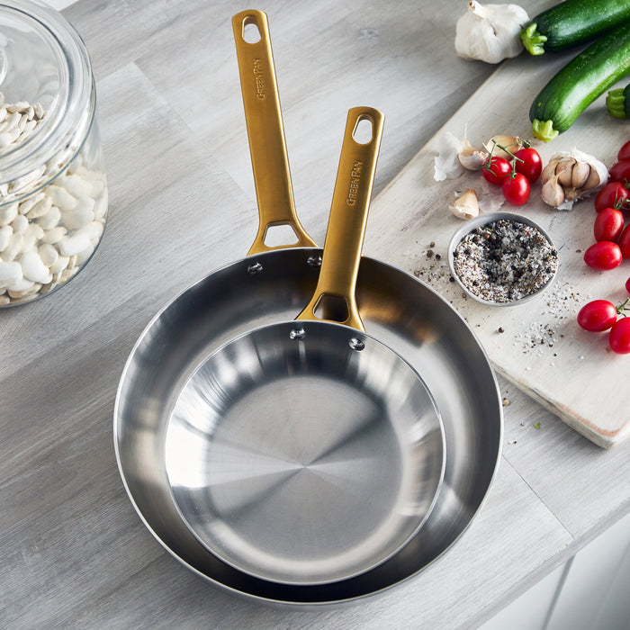 Stainless steel frying pans with gold handles on a kitchen counter with vegetables and spices.