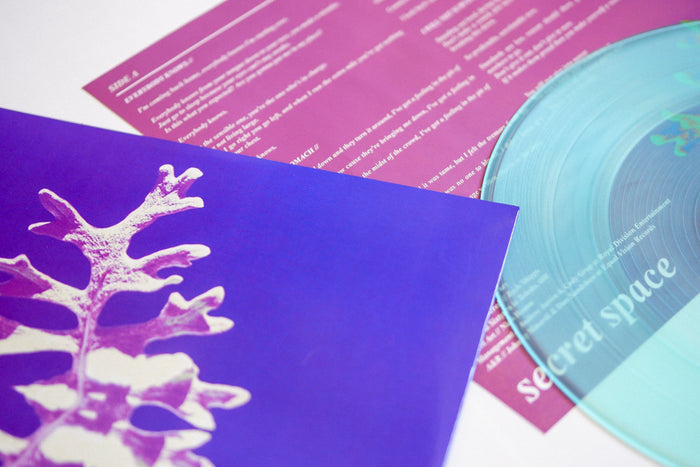 A limited edition Secret Space vinyl in Coke Bottle Green is shown beside a purple sleeve with a white leaf and a pink insert with white text.