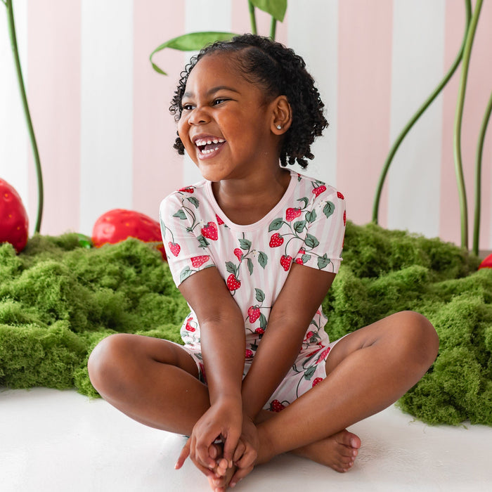 Smiling girl sitting on the floor in front of a strawberry backdrop wearing the Short Sleeve Pajamas in Strawberry Fields