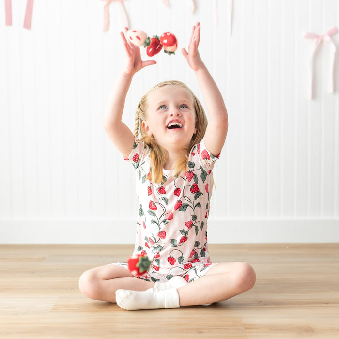 Young girl sitting cross legged on the floor wearing the Short Sleeve Pajamas in Strawberry Fields throwing plush toy strawberries in the air