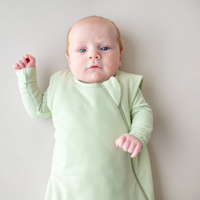 Baby wearing a light green sleep bag and footie against a plain background