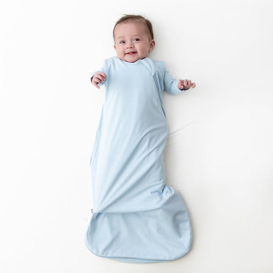 Smiling infant laying on a white background wearing the Sleep Bag in Breeze 0.5 with a matching romper on underneath