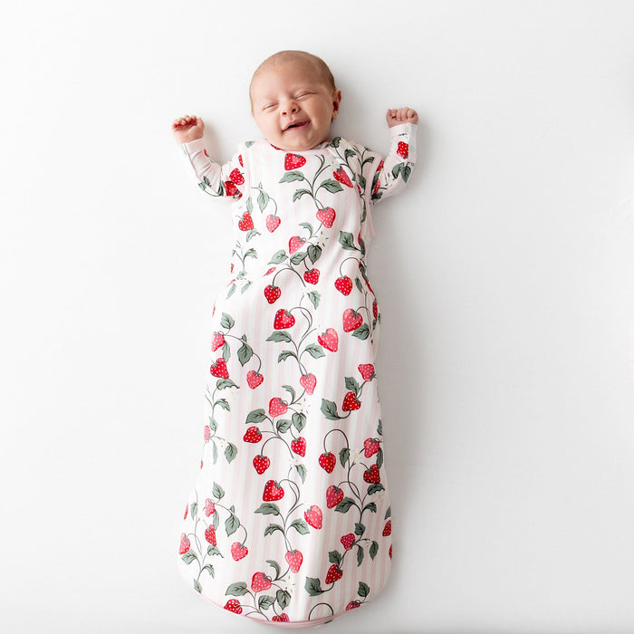 Newborn smiling laying on a light pink surface wearing the Sleep Bag in Strawberry Fields 1.0 with matching romper on underneath