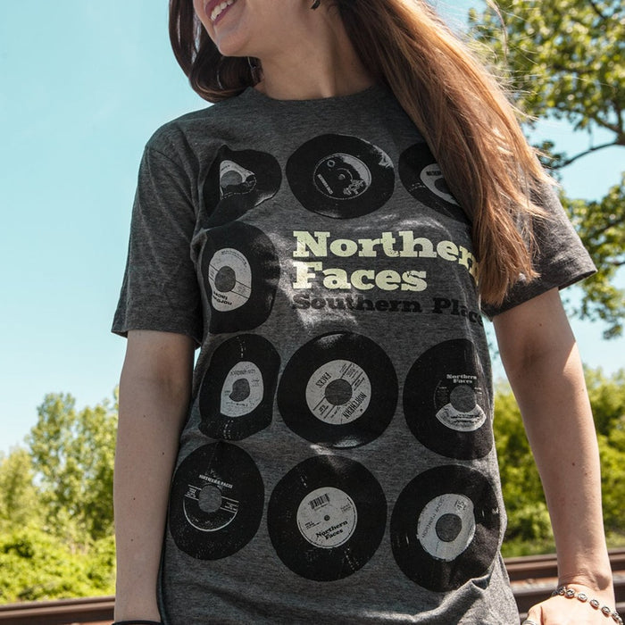 A woman stands outdoors in a Northern Faces Heather Charcoal T-shirt with vinyl records and the phrase “Northern Faces Southern Place.” Behind her are trees and a blue sky.