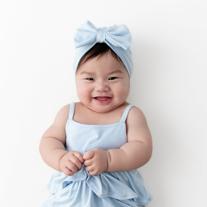Smiling infant wearing the Bow Headwrap in Breeze with matching spaghetti strap ruffle romper