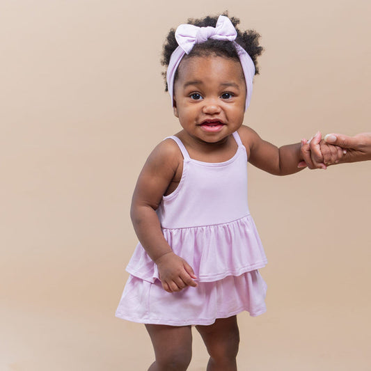 Young toddler standing wearing the Spaghetti Strap Ruffle Romper in Thistle with matching bow holding mother's hand