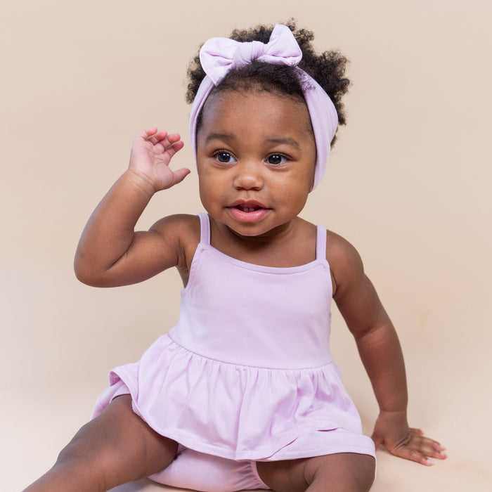 Young toddler sitting on the floor wearing the Spaghetti Strap Ruffle Romper in Thistle with matching bow