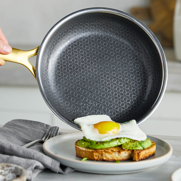 Gold ceramic nonstick frypan held over a plate with avocado toast and a sunny-side-up egg.