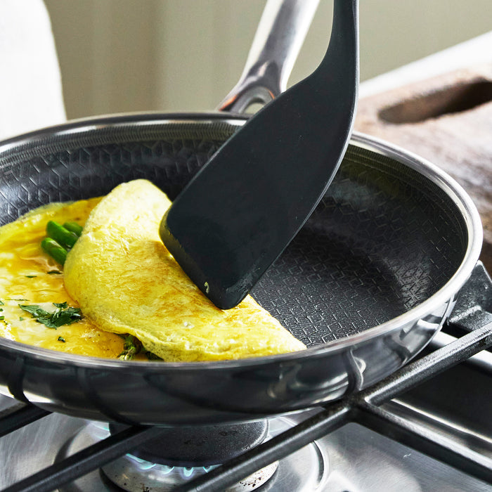Omelet with vegetables being folded with a black spatula in a white Spectra ceramic nonstick frypan on a stove.