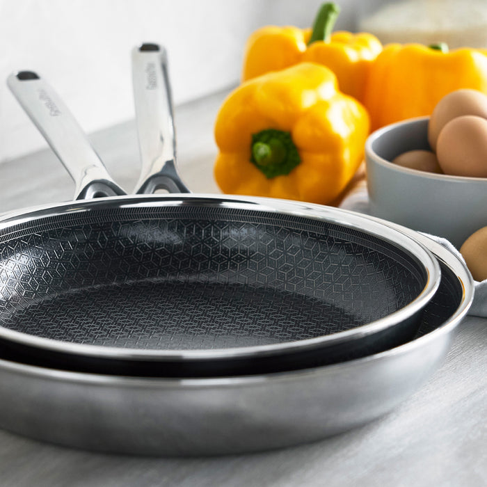 Set of two grey ceramic frypans on a kitchen counter with yellow peppers and eggs nearby.