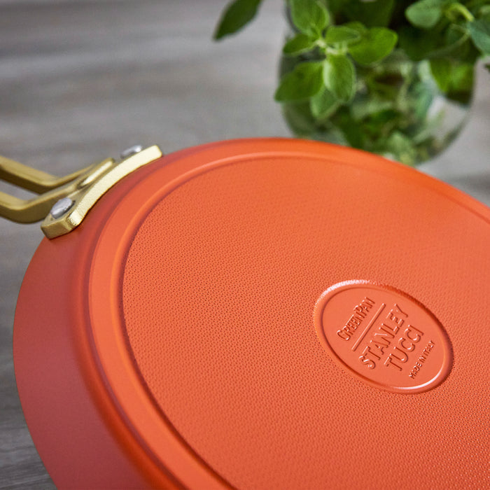 Ceramic nonstick 10-inch pan in orange, next to a green potted plant on a wooden surface.